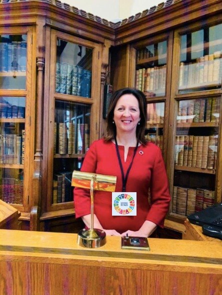 Jacqueline Winstanley in a red dress with a UN Sustainable Development Goals badge, in a wood panelled library at Windsor, November 2019