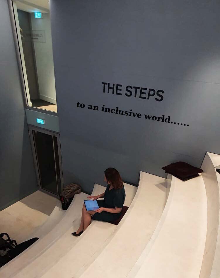 Jacqueline sitting with a tablet on wide white steps, beneath a wall that reads: The steps to an inclusive world